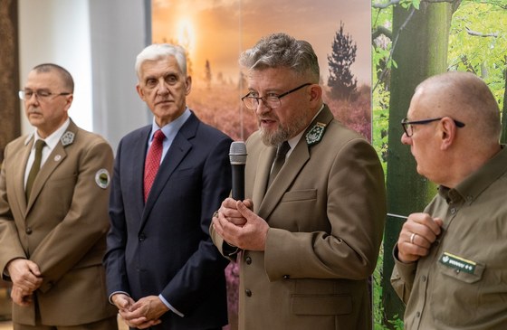 Na tle fotościanki z motywem leśnym stoi czterech mężczyzn. Jeden z nich trzyma mikrofon i przemawia.