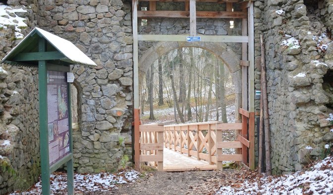 Na fotografii jest widoczna zbudowana z kamienia brama, w dalszym planie widać drewnianą kładkę/  Fot. M. Majchrzyk