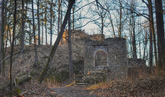 Na zdjęciu są widoczne ruiny zamku oraz fragmenty drewnianej, zniszczonej kładki
