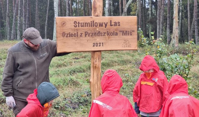 Zdjęcie przedstawia leśnika wraz z dziećmi przy pamiątkowej tablicy o treści "Stumilowy Las Dzieci z Przedszkola "Miś" 2021