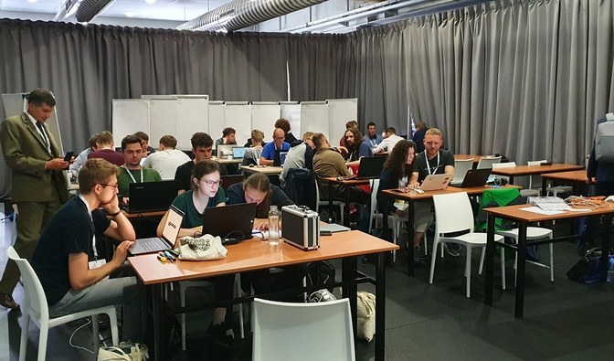 Grupa ludzi siedząca przy stołach i pracująca na laptopach. Przy jednym stole stoi leśnik i przygląda się pracującym programistom. Fot. Kamil Onoszko