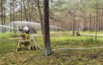 Na fotografii są widoczne osoby w strojach straży pożarnej, które rozlewają wodę w lesie