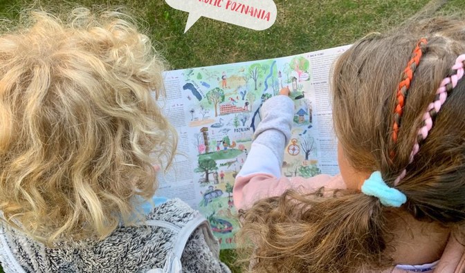 Na zdjęciu ujęcie od góry dziewczynki i chłopca, którzy oglądają NIEMAPĘ Lasów Okolic Poznania. Dziewczynka coś wskazuje palcem na mapie, a przy chłopcu jest rysunkowy dymek z tekstem "Lasy okolic Poznania".