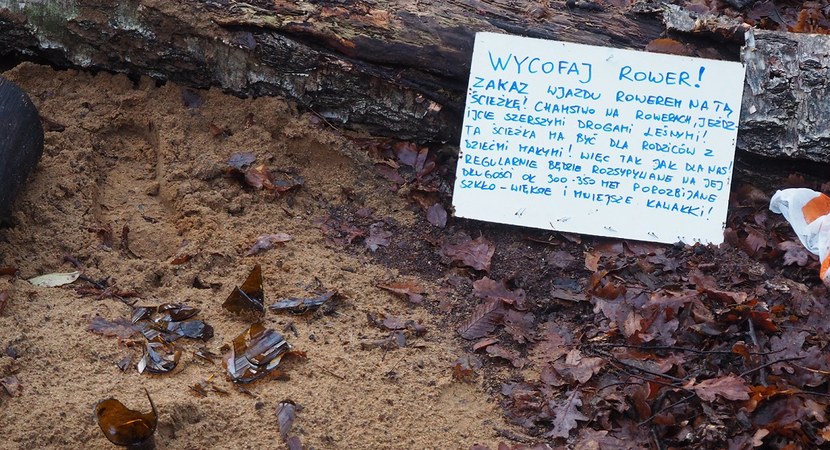 Na zdjęciu widać szkło z rozbitych butelek, ułożone na ścieżce w lesie, o gałęzie leży oparta kartka z ostrzeżeniem