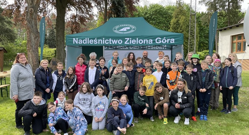 Na tle zielonego namiotu z logo Lasów Państwowych i napisem "Nadleśnictwo Zielona Góra" grupa dzieci z nauczycielkami i leśnikiem.