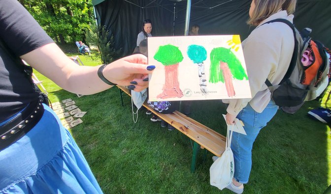 Kobieca dłoń trzymająca rysunek farbkami. Na rysunku drzewa i logo Lasów Państwowych.