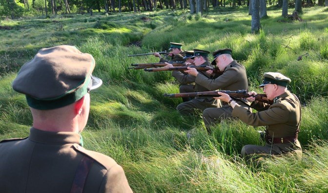 Mężczyźni w mundurach z epoki, kucają w okopie. W ręku trzymają broń przygotowana do strzału.