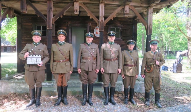 Przed drewnianym domem, przy którego wejściu na ścianie wisi godło Polski, stoją mężczyźni w leśnych mundurach z epoki. Jeden z nich trzyma klaps filmowy.