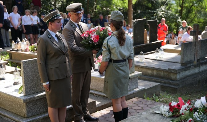 Kobieta i mężczyzna w mundurze leśnika, stoją z kwiatami na cmentarzu. Przed nimi harcerka.