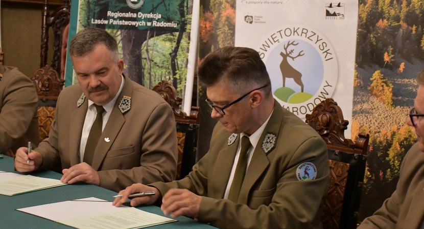 Przy stole mężczyźni w mundurach leśnika i pracownika parku narodowego podpisuję porozumienie. W tle roll-upy z logo Lasów Państwowych i Świętokrzyskiego Parku Narodowego.