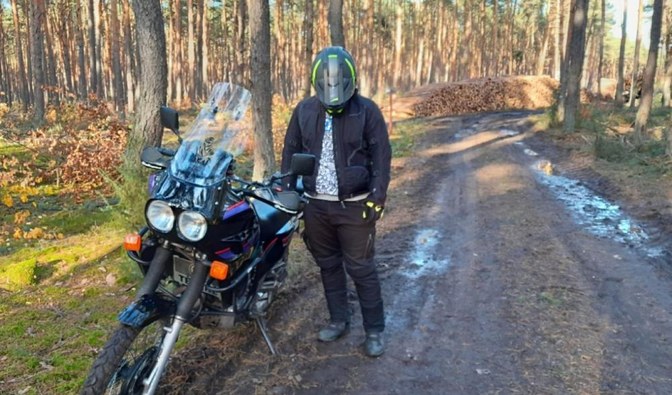 Mężczyzna w kasku motocyklowym stojący przy motorze, zatrzymany na drodze leśnej
